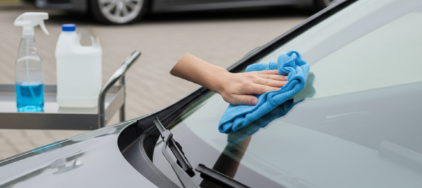 Een hand reinigt de voorruit van een geparkeerde auto met een microvezeldoek, naast producten voor ruitenonderhoud.